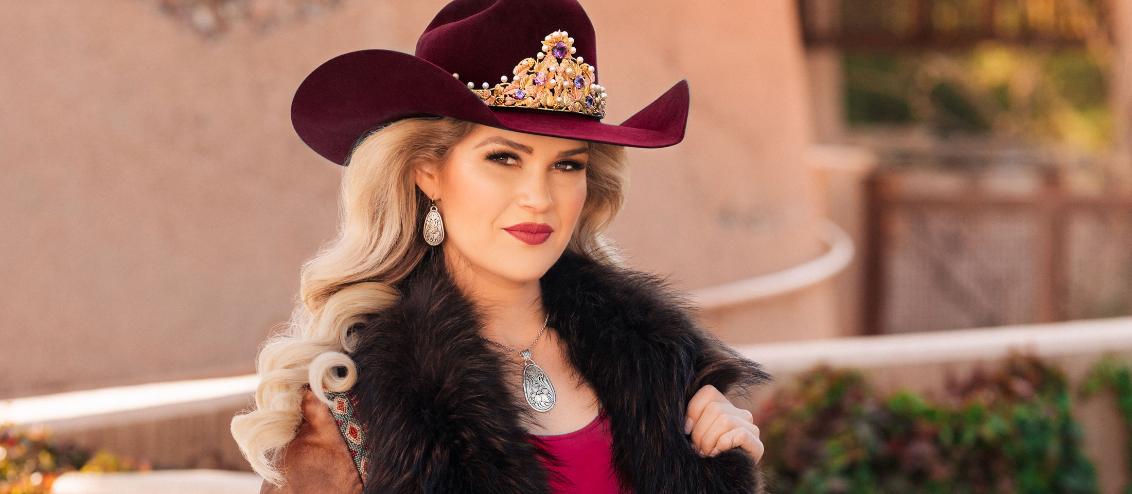 Miss Rodeo America is wearing a brown jacket with black fur around the neck/ collar and a maroon hat, posing for the camera.